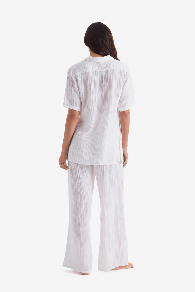Woman turned away wearing a white short sleeve button up and white beach pants